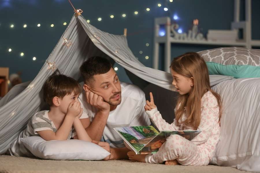 Un père et ses deux enfants lisent un livre sous une cabane en drap illuminée, parfait pour générer des histoires pour enfants avec l'IA.